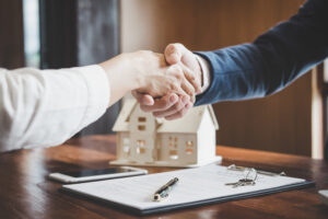 Two people shake hands over a miniature house on a desk, with a smartphone and documents nearby.