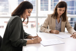 Two women sign documents at a bright office desk, one in a dark green blouse and the other in a beige blazer.